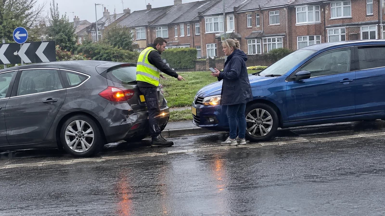 Two drivers arguing at the kerb after a minor bump at a suburban roundabout in the rain